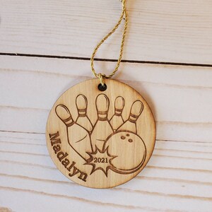 May include: A round wooden ornament with a bowling ball and pins design. The ornament is personalized with the name "Madalyn" and the year "2021".