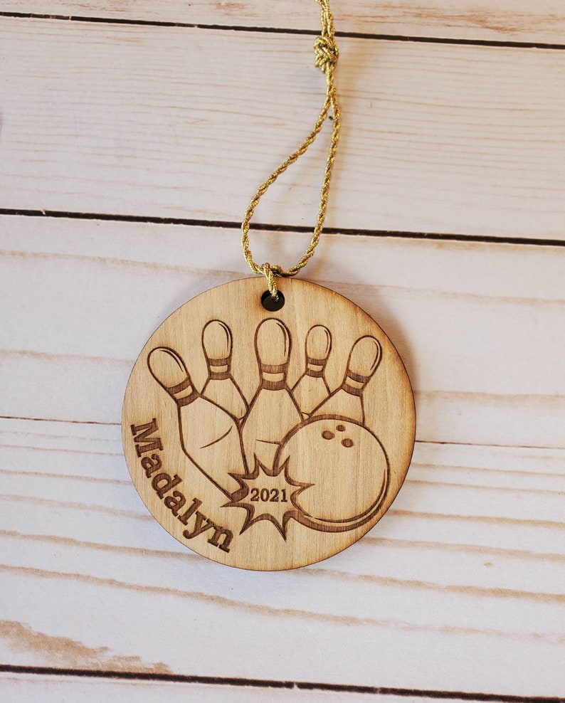 May include: A round wooden ornament with a bowling ball and pins design. The ornament is personalized with the name "Madalyn" and the year "2021".
