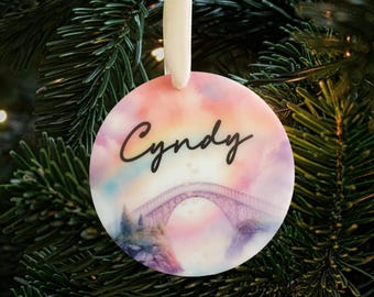 Puede incluir: Un adorno navideño redondo y blanco con un diseño de acuarela de un puente y nubes. El adorno tiene el nombre "Cindy" escrito en cursiva negra.