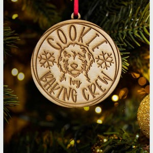 May include: Wooden Christmas ornament with the text "COOKIE BAKING CREW" and a dog illustration. The ornament is round with snowflake details and a red ribbon for hanging. The word "Ivy" is also on the ornament.