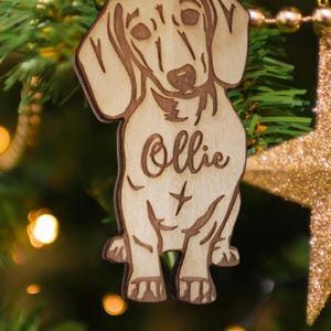 May include: Wooden Christmas ornament shaped like a dachshund dog with the name "Ollie" engraved on it. The ornament is brown and has a light wood grain.