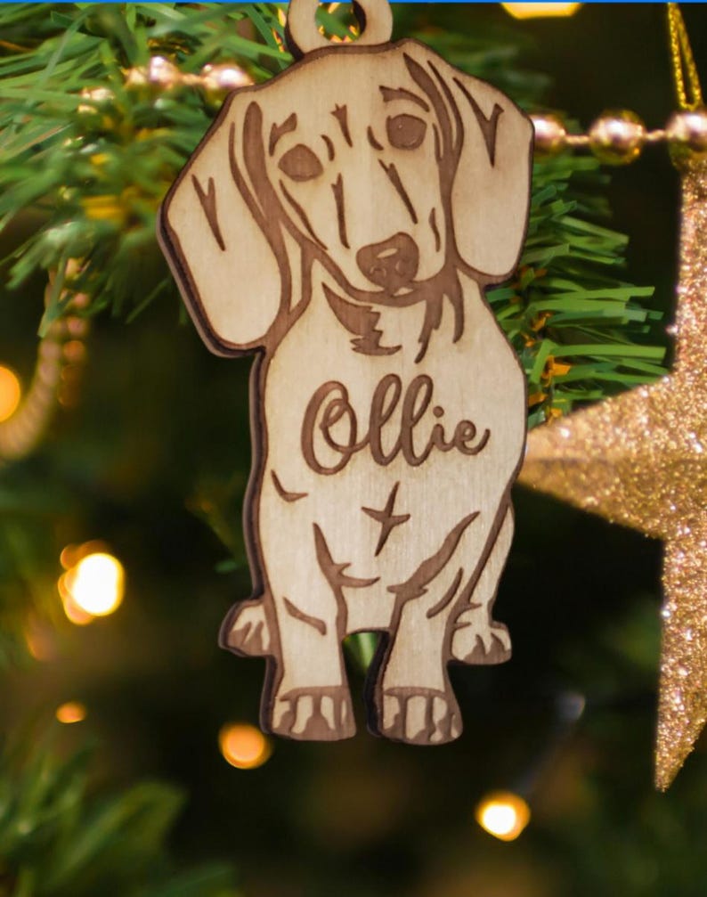 May include: Wooden Christmas ornament shaped like a dachshund dog with the name "Ollie" engraved on it. The ornament is brown and has a light wood grain.