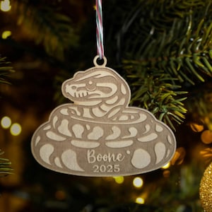 May include: Wooden Christmas ornament shaped like a coiled snake, with the name "Boone" and the year "2025" engraved on it. The ornament is hanging from a red, green, and white string against a blurred Christmas tree background.
