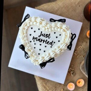 May include: A heart-shaped cake with white frosting, decorated with black bows and small black hearts. The words "Just married" are written in black script on the cake. The cake sits on a white surface.