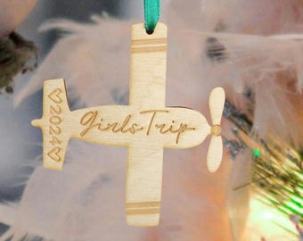 Puede incluir: Adorno de avión de madera con el texto "Girls Trip" y el año "2024" en forma de corazón. El adorno está pintado en un color madera natural y tiene una cinta verde.