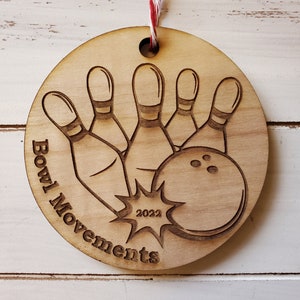 May include: A wooden ornament with a bowling ball and pins design. The ornament is brown and has the text "Bowl Movements 2022" on it.