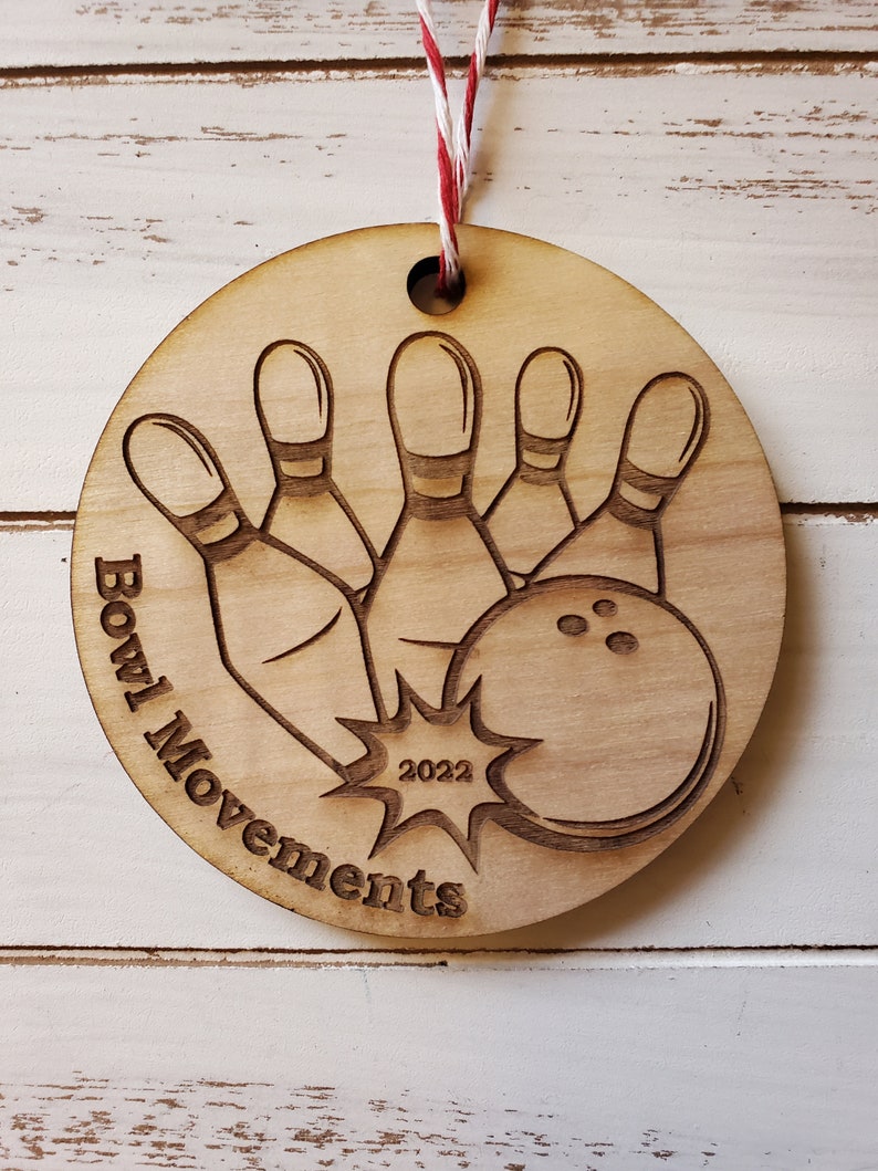 May include: A wooden ornament with a bowling ball and pins design. The ornament is brown and has the text "Bowl Movements 2022" on it.