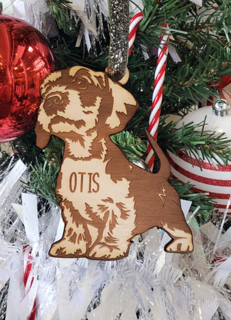 May include: A wooden Christmas ornament shaped like a dachshund dog with the name "Otis" engraved on it. The ornament is hanging on a Christmas tree with red and white candy canes and other ornaments.