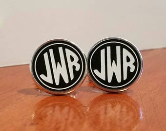 May include: A pair of round cufflinks with a black background and the white letters "JWB" in a bold, sans-serif font. The cufflinks have a silver-colored border and are displayed on a wooden surface.