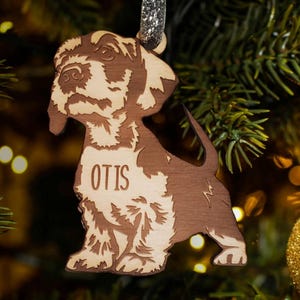 May include: A wooden Christmas ornament shaped like a dog with the name "Otis" engraved on it. The ornament is brown and has a glittery silver ribbon attached to the top.