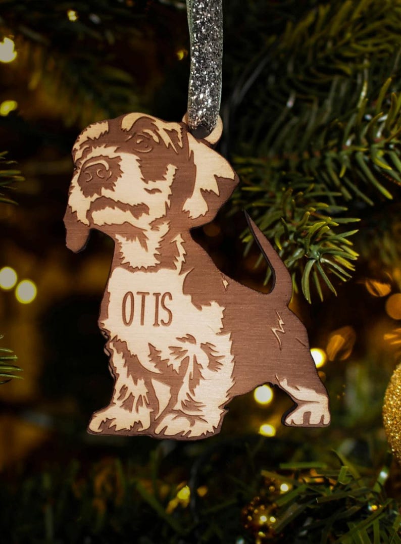 May include: A wooden Christmas ornament shaped like a dog with the name "Otis" engraved on it. The ornament is brown and has a glittery silver ribbon attached to the top.