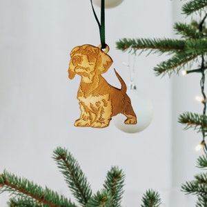 May include: A wooden Christmas ornament shaped like a brown and white puppy dog. The ornament is hanging from a green ribbon on a Christmas tree.