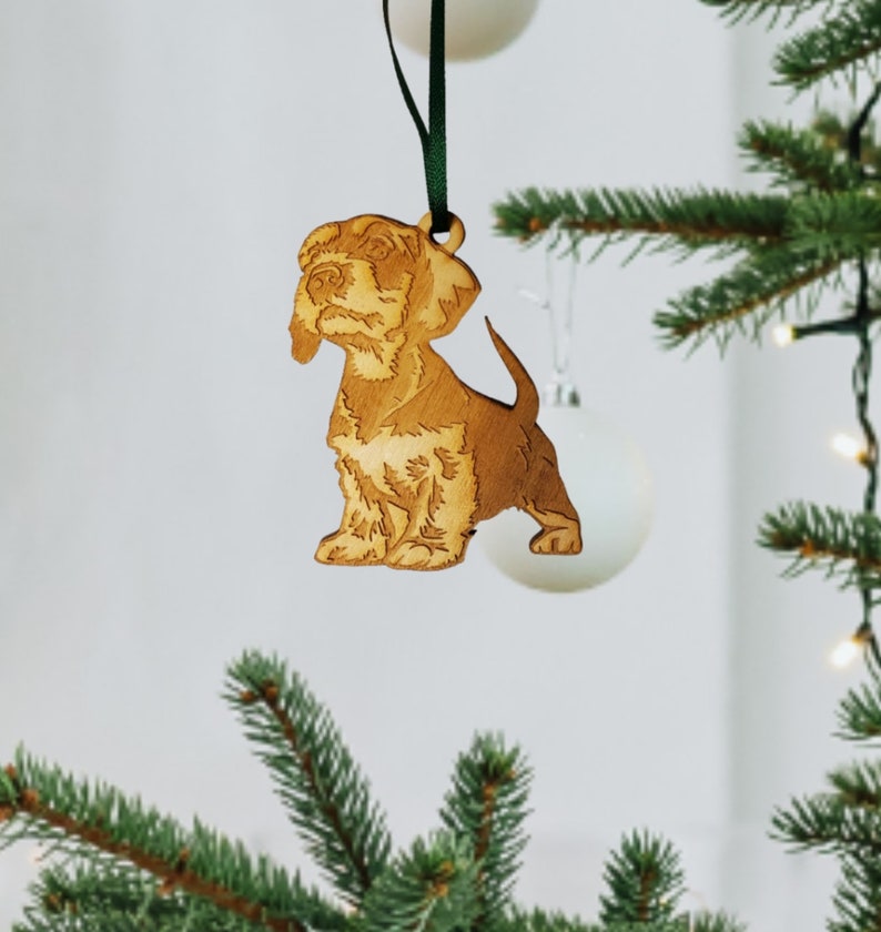 May include: A wooden Christmas ornament shaped like a brown and white puppy dog. The ornament is hanging from a green ribbon on a Christmas tree.