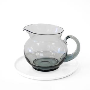 Puede incluir: Una jarra de vidrio transparente con un tinte gris y un cuerpo redondeado. La jarra tiene un asa y se asienta sobre un platillo blanco.