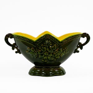 May include: A green ceramic bowl with yellow accents and ornate handles. The bowl has a floral design and a raised base.