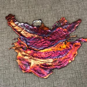 May include: An irregular, flat metal pendant featuring a vibrant, iridescent finish with swirling gradients of metallic copper, deep magenta, and violet. The surface has a textured, molten appearance with a small loop at the top for hanging.