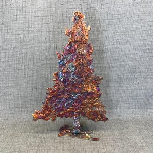 Peut inclure: Une sculpture artisanale d'arbre de Noël en métal coloré. L'arbre présente une surface texturée avec des nuances d'orange, de rouge, de violet et de bleu. L'arbre est sur une petite base, et le design général est artistique et décoratif.