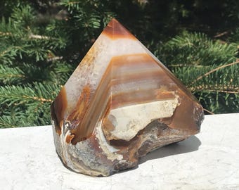 Pode incluir: Uma pedra de ágata polida em forma de pirâmide, com camadas de tons castanhos, brancos e laranja. A base da pedra natural é áspera e texturizada, enquanto o topo é liso e pontiagudo. O fundo apresenta folhagem verde.