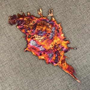 May include: Abstract metal art piece with a fiery color palette. The artwork features a blend of orange, red, purple, and white hues, creating a dynamic and textured appearance. The piece has an irregular, organic shape, suggesting a unique, handcrafted design.