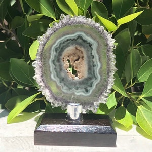 May include: A green and purple agate geode slice with a crystal edge, mounted on a black wooden base with a silver metal stand.