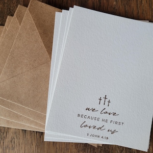 May include: A stack of blank white cards with brown envelopes. The cards have a simple design with the text "we love because he first loved us 1 John 4:19" printed in brown ink.