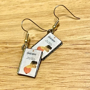 Op de afbeelding: Gouden oorbellen met een miniatuur boekbedel. De boekomslag is wit met gouden letters die "Shakespeare The Taming of the Shrew" lezen.