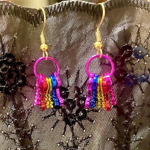 May include: A pair of handmade earrings featuring a gold-colored hook and a small, bright pink hoop. Attached to the hoop are several small, colorful, metallic rectangles in a rainbow pattern. The earrings are set against a black, sequined background.