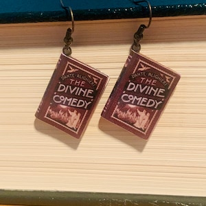 May include: A pair of earrings featuring miniature book charms with the title "The Divine Comedy" by Dante Alighieri. The charms are brown with gold accents and are hanging from gold hooks.