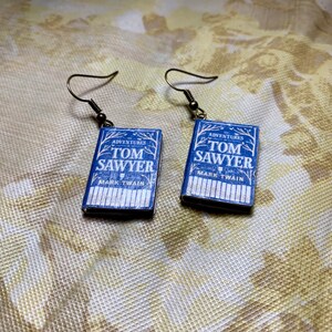 May include: A pair of blue book-shaped earrings with the title "The Adventures of Tom Sawyer" by Mark Twain. The earrings have gold hooks.