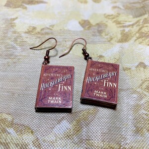 May include: A pair of earrings featuring miniature book charms. Each charm is a replica of "The Adventures of Huckleberry Finn" by Mark Twain, with a brown cover and white lettering. The earrings have copper-colored hooks.
