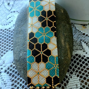 Puede incluir: Una pulsera de cuentas con un patr&oacute;n floral geom&eacute;trico en negro, blanco y turquesa. La pulsera tiene un cierre dorado y detalles dorados. La pulsera mide aproximadamente 3,8 cm de ancho.