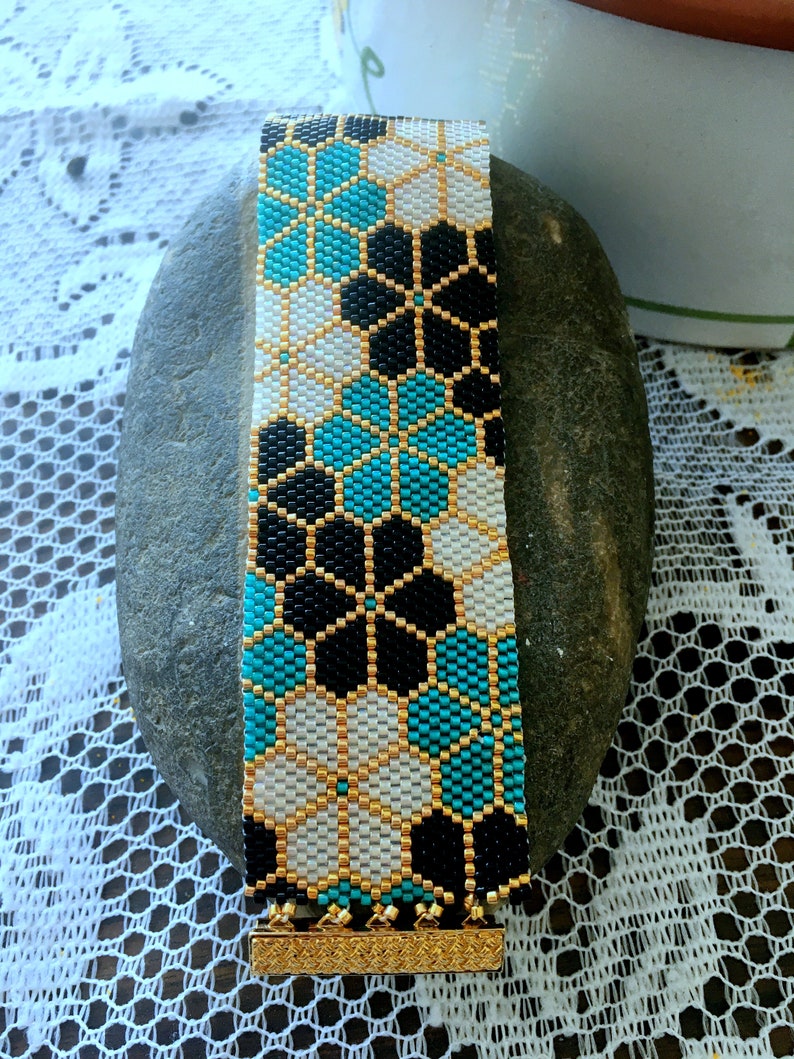 Puede incluir: Una pulsera de cuentas con un patr&oacute;n floral geom&eacute;trico en negro, blanco y turquesa. La pulsera tiene un cierre dorado y detalles dorados. La pulsera mide aproximadamente 3,8 cm de ancho.