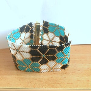 Puede incluir: Una pulsera ancha de cuentas con un patr&oacute;n geom&eacute;trico. La pulsera presenta un dise&ntilde;o repetido de cuentas negras, blancas y turquesas, acentuadas con cuentas doradas. La pulsera se asegura con un cierre dorado.