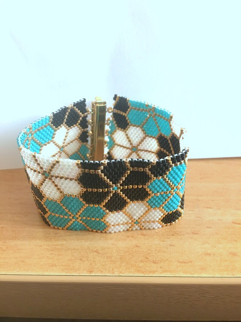 Puede incluir: Una pulsera ancha de cuentas con un patr&oacute;n geom&eacute;trico. La pulsera presenta un dise&ntilde;o repetido de cuentas negras, blancas y turquesas, acentuadas con cuentas doradas. La pulsera se asegura con un cierre dorado.