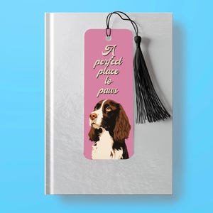 May include: Pink bookmark featuring a brown and white dog illustration and the text "A perfect place to paws". The bookmark has a black tassel.