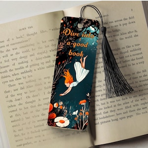 May include: A bookmark with a dark blue background and a girl in a white dress falling into a book. The bookmark has the text "Dive into a good book" in gold lettering. The bookmark has a black tassel.