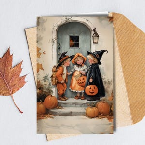 May include: A watercolour illustration shows three children in Halloween costumes on a porch with pumpkins. One child is in a witch costume, another in a pumpkin costume, and the third in a pilgrim costume. The image is on a greeting card with a brown envelope and a fall leaf.