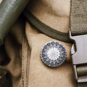 May include: A close-up of a brown canvas backpack with a green strap and a silver pin with a geometric design.