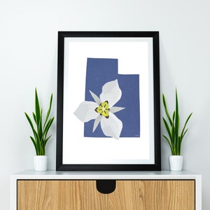May include: A framed print of the state of Utah featuring a white sego lily flower on a blue background.