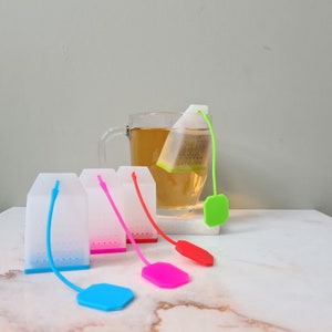 Peut inclure: Quatre infuseurs à thé en silicone blanc avec des cordons de différentes couleurs. Les cordons sont bleu, rose, rouge et vert. Les infuseurs sont sur une surface blanche. Une tasse en verre avec du thé est en arrière-plan.