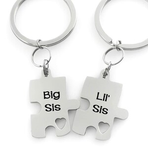 Può includere: Due portachiavi a forma di puzzle argentati. Uno recita "Big Sis" e l'altro "Lil' Sis". Ogni pezzo ha un ritaglio a forma di cuore. I portachiavi hanno un anello argentato per attaccare le chiavi.
