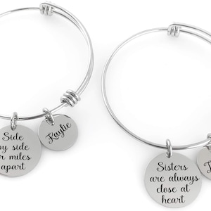 Può includere: Due braccialetti a cerchio regolabili color argento. Ogni braccialetto ha un ciondolo rotondo con un nome e un ciondolo più grande con una citazione ispiratrice. Uno dice "Side by side or miles apart" e l'altro "Sisters are always close at heart."