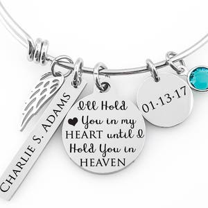 Puede incluir: Una pulsera de brazalete plateada con múltiples dijes. Los dijes incluyen un ala, una etiqueta rectangular con "CHARLIE S. ADAMS", un dije redondo con "I'll Hold You in my HEART until I Hold You in HEAVEN", un dije con la fecha "01-13-17" y un cristal turquesa.