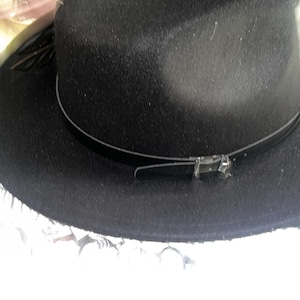 May include: A black fedora hat with a textured finish. The hat features a thin, dark band around the crown and a decorative buckle. The brim is slightly upturned. A stylish accessory for any wardrobe.