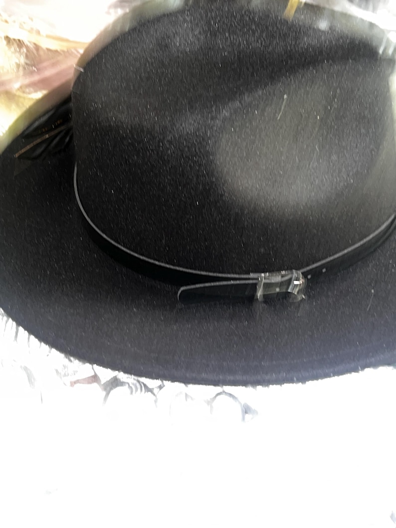 May include: A black fedora hat with a textured finish. The hat features a thin, dark band around the crown and a decorative buckle. The brim is slightly upturned. A stylish accessory for any wardrobe.