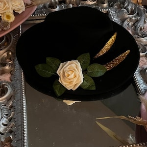 May include: Black hat with a cream-colored rose and green leaves. Two gold glitter accents are on the hat. The hat is sitting on a decorative mirror.
