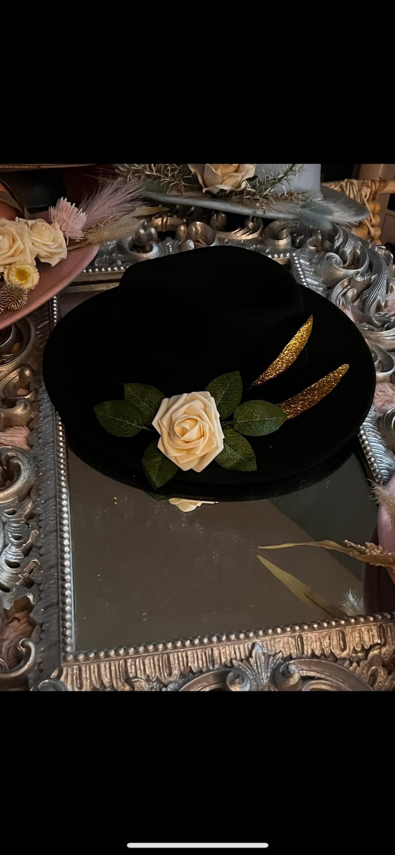 May include: Black hat with a cream-colored rose and green leaves. Two gold glitter accents are on the hat. The hat is sitting on a decorative mirror.