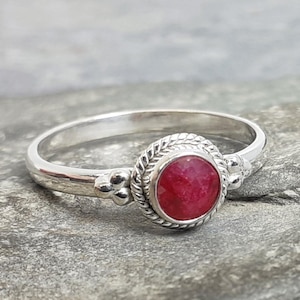 Może przedstawiać: Srebrny pierścionek z czerwonym kamieniem szlachetnym osadzonym w oprawie typu bezel. Pierścionek ma zdobiony pasek z koralikami.