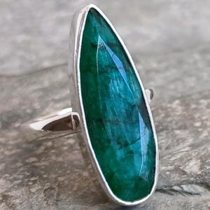 May include: A sterling silver ring featuring a large, emerald-cut, deep green gemstone. The gemstone is set in a polished silver bezel, creating a striking contrast. The ring's band is simple and elegant, highlighting the vibrant green stone.