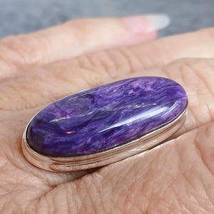 Puede incluir: Cabujón ovalado de piedra preciosa de charoíta púrpura engastado en un bisel plateado. La piedra muestra patrones arremolinados de tonos lavanda y violeta. Una pieza de joyería, posiblemente un anillo o colgante.